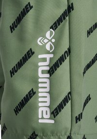 Grön atletisk tyg med ett upprepande svart "HUMMEL"-logotyp och en vit logotypaccent. Slät textur, modern design och livlig färgkontrast.