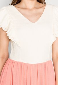 Donna che indossa un vestito con una parte superiore bianca texturizzata, con maniche corte arricciate e una gonna rosa plissè.
