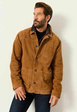 Mann mit Bart, der eine braune Wildlederjacke über einem karierten Hemd mit Kragen und dunklen Hosen trägt, schaut nach links vor einfachem Hintergrund.