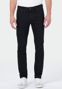 Homme portant un jean slim noir et des baskets blanches, se tenant devant un fond clair uni.