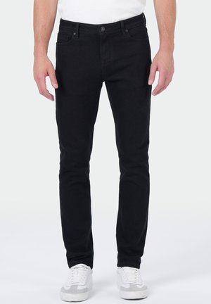 Man in slim-fit zwarte jeans en witte sneakers, staande tegen een effen lichte achtergrond.