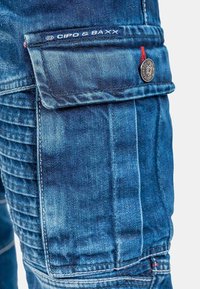 Pantalons cargo en denim bleu avec des coutures détaillées, présentant une poche rabat, une étiquette de marque et un accent de bouton métallique.
