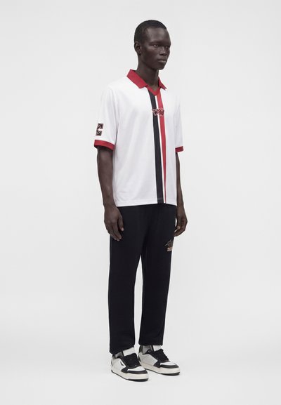 Homme debout portant un polo blanc à rayures verticales rouges et noires, pantalon noir et baskets blanches et noires sur fond uni.