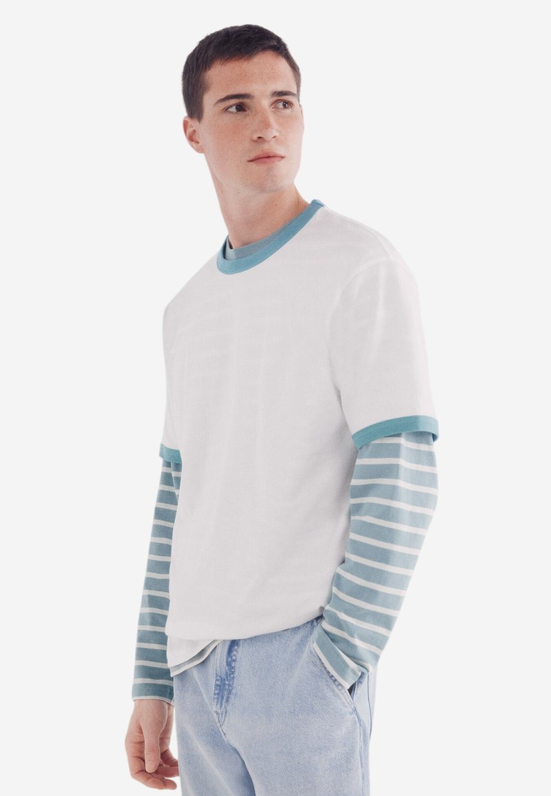 Weißes T-Shirt mit kurzen Ärmeln und tealfarbenem Rundhalsausschnitt, über einem langärmligen, hellblauen und weißen gestreiften Hemd getragen, kombiniert mit hellblauen Jeans.