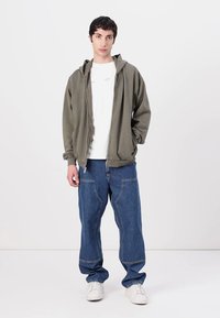 Olive green zip-up hoodie, white t-shirt, blue denim pants, and white sneakers. Casual streetwear style with relaxed fit and simple design.