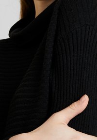 Černý žebrovaný pletený svetr s vysokým přeloženým límcem. Textura je měkká a hrubá, celkově má přiléhavý střih.