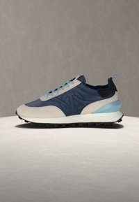 Baskets bleu et beige avec lacets bleu clair et accent au talon, semelle noire, et motif "V" en relief sur le côté, posées sur une surface grise lisse.