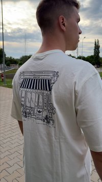 Jaunas vyras, vilkintis baltą marškinėlį su juodu piešiniu, vaizduojančiu kavinės fasadą su dryžuota skėčiu ir lauko kėdėmis, stovi lauke šalia šaligatvio.