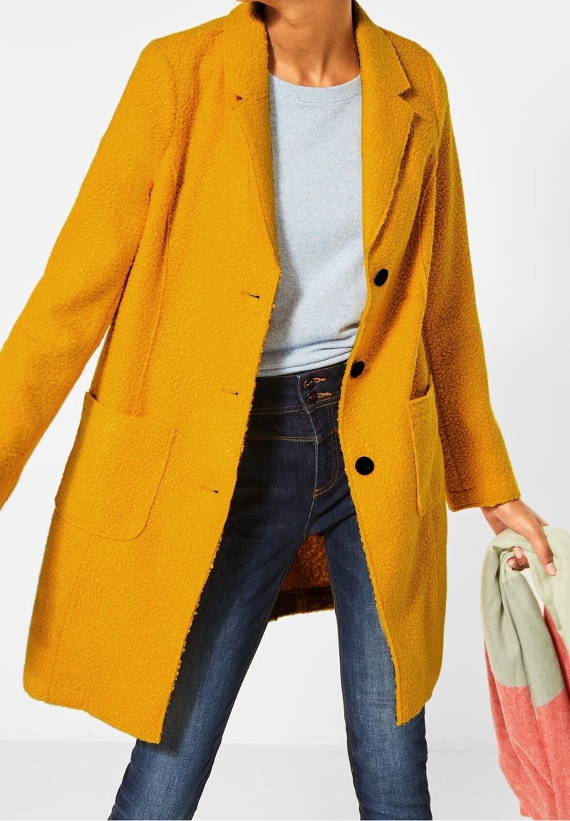 Femme portant un manteau texturé jaune moutarde par-dessus une chemise gris clair et un jean bleu foncé, tenant un tissu plié rose et vert à la main.