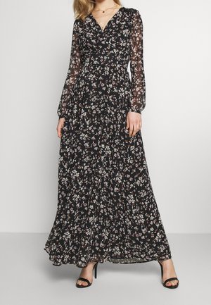 Mujer con un vestido largo negro con estampado floral, mangas largas transparentes y sandalias negras de tacón alto, de pie contra un fondo liso.