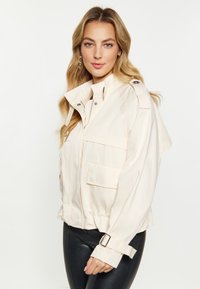 Veste en coton beige clair avec un col rabattu, fermeture éclair à l'avant, poches latérales et poignets réglables. Présente une coupe décontractée et des pattes d'épaule.
