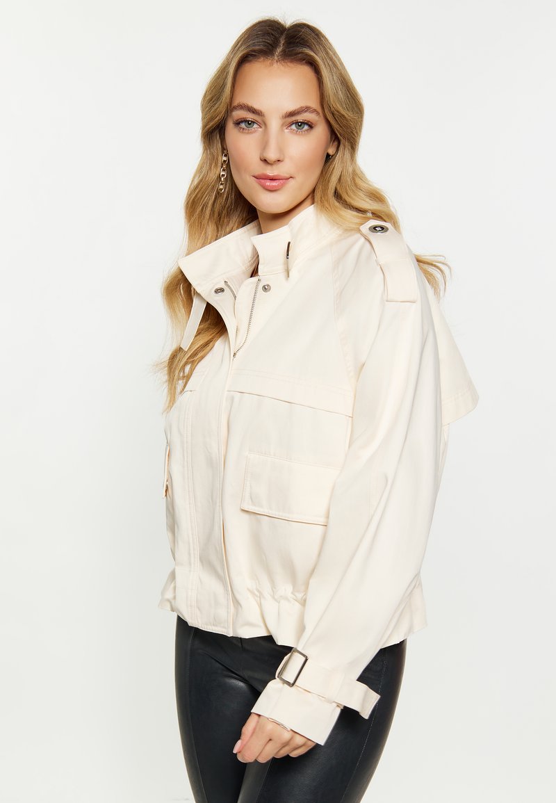 Veste en coton beige clair avec un col rabattu, fermeture éclair à l'avant, poches latérales et poignets réglables. Présente une coupe décontractée et des pattes d'épaule.