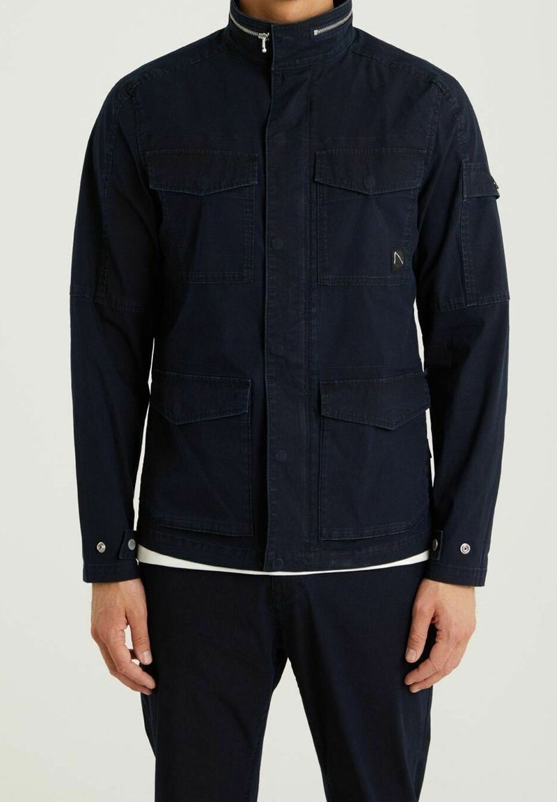 Navy cotton jacket with a stand collar, front button closure, and multiple pockets. Features textured fabric and subtle hardware accents.