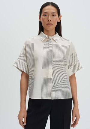 Frau mit langen dunklen Haaren, die eine kurzärmelige, cremefarbene Bluse mit schwarzen vertikalen und diagonalen Streifen trägt, kombiniert mit schwarzen Hosen.