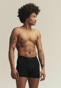 Bóxer negros hechos de tela elástica, con un ajuste ceñido y una cinturilla elástica. El modelo tiene el pelo corto y rizado y tatuajes visibles.