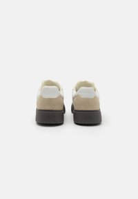 White and beige sneakers with a suede heel and black rubber sole. Features a minimalistic design with rounded toe and stitched accents.