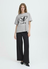 Camiseta gris de manga corta con un gráfico de águila negra y el texto "NACIDO PARA SER SALVAJE", combinada con pantalones anchos negros y zapatos negros de punta.