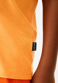 Orange bomulds-T-shirt med en let glans, der har et sort mærke med teksten "BAKER" nær venstre søm. Glat tekstur, korte ærmer.