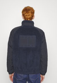 Giacca blu navy con corpo in pile e maniche trapuntate. Presenta un logo "TOMMY JEANS" su una toppa rettangolare sul retro.