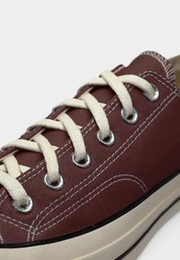 Μπορντό πάνινο αθλητικό παπούτσι με λευκή ραφή, κορδόνια σε μπεζ χρώμα περασμένα από ασημένια πριντζάκια και λευκή λαστιχένια σόλα με μαύρο περίγραμμα.