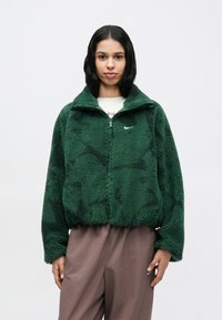 Ζακέτα από φλις σε σκούρο πράσινο με υφή, φερμουάρ μπροστά, όρθιο γιακά και διακριτικό λογότυπο στο αριστερό στήθος. Κοντό μήκος.