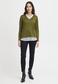 Zielony, fakturowany sweter nałożony na czarną i białą koszulę w kratę, w połączeniu z czarnymi spodniami typu slim fit i czarnymi botkami za kostkę.