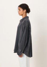 Jauna moteris su garbanotais plaukais, surištais žemu kuodu, dėvi laisvą tamsiai pilkos spalvos kordurovinę palaidinę ir šviesiai mėlynas džinsines kelnes, stovinti šonu prieš baltą foną.