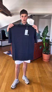 Camiseta negra de manga corta sostenida por una persona, con un pequeño logo y una etiqueta de precio adjunta. El fondo incluye una planta y muebles.