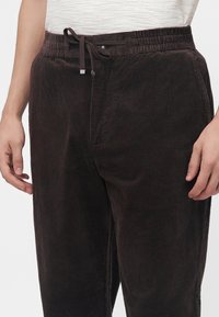 Gros plan d’une personne portant un pantalon en velours côtelé marron foncé avec une taille élastique et un cordon, et une chemise blanche rentrée.
