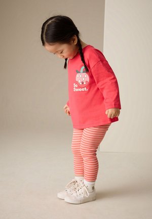 Niño pequeño con el pelo trenzado, que lleva una camiseta roja de manga larga, leggings a rayas y zapatillas blancas, mirando hacia abajo mientras está de pie.