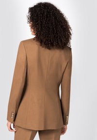 Bordeaux skräddarsydd kavaj med en åtsittande design, som har en mjuk textur och axelkuddar. Knappdetaljer är synliga på ärmarna.