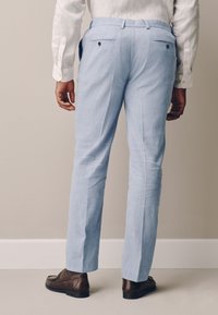 Man wearing light blue trousers, white long-sleeve shirt, brown loafers, standing indoors against plain beige wall and white baseboard.