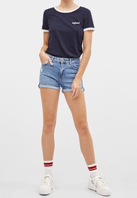 Camiseta de marina con ribete blanco y texto "¡super!", combinada con pantalones cortos de mezclilla enrollados. Zapatillas blancas con calcetines de rayas rojas completan el look.