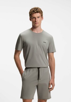 Homme aux cheveux bouclés portant une chemise gris clair à manches courtes et un short assorti avec un cordon, debout devant un fond blanc uni.