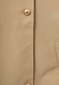 Beige stof med en glat tekstur, der har en stor metalknap og sidelomme. Sømme detaljer er synlige langs forsiden.