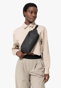 Borsa a tracolla compatta nera con forma arrotondata, tracolla regolabile e chiusura a zip. Indossata sopra una camicia a righe beige e pantaloni grigi.