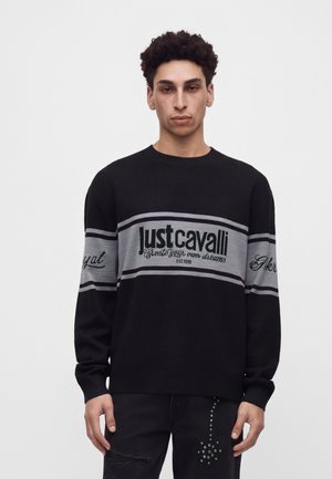 Jeune homme aux cheveux bouclés portant un pull Just Cavalli noir et gris avec un texte et un pantalon noir à clous.