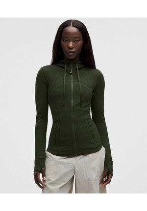 Mujer con una sudadera ajustada verde oscuro con cremallera y agujeros para los pulgares y pantalones beige, de pie frente a un fondo liso claro.