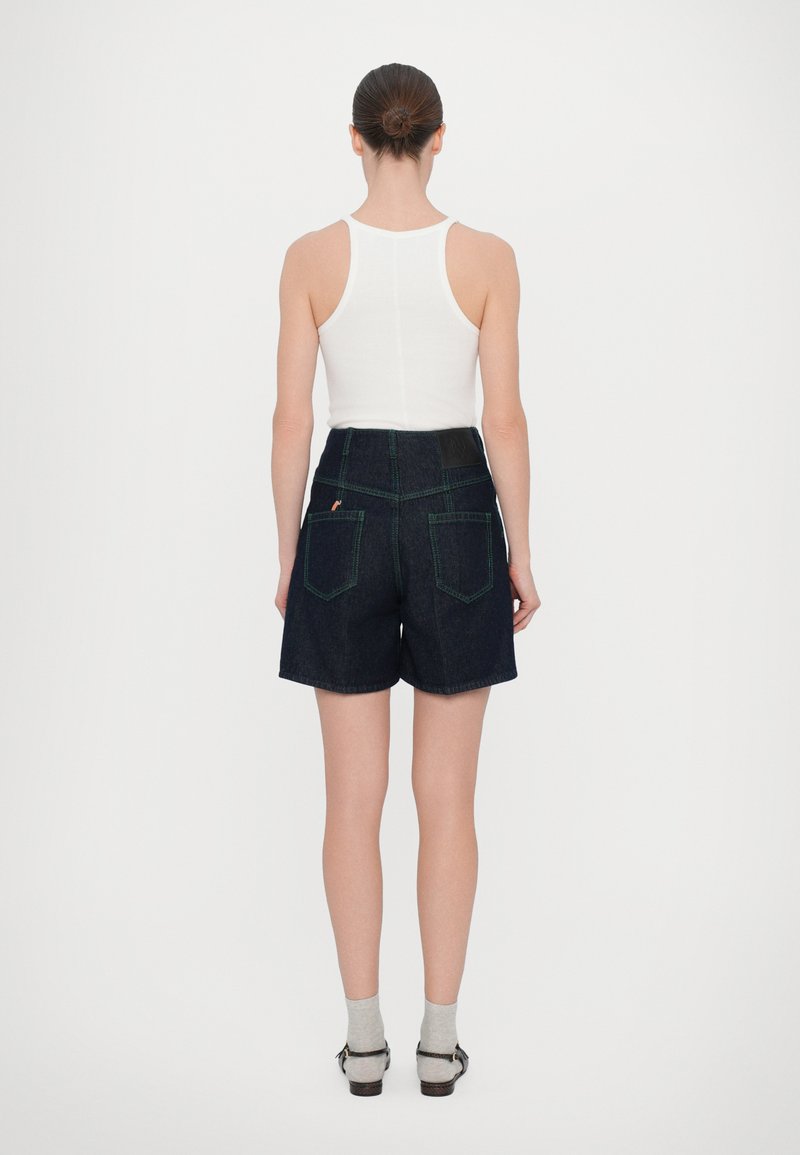 Denim-Shorts in dunkler Waschung mit grünen Nähten. Hohe Taille, zwei Gesäßtaschen und ein glatter Bund. Das Model trägt ein weißes Tanktop.