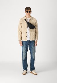 Beige katoenen jack met zwarte knopen, witte t-shirt, blauwe spijkerbroek, zwarte schoudertas en witte sneakers met bruine zolen. Zwart zonnebril.