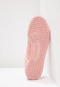 Semelle de basket Reebok rose avec un motif de bande de roulement en forme de diamants et de cercles, appuyée contre un mur d’angle blanc sur une surface blanche.