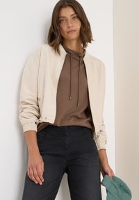 Beige bomberjack met elastische manchetten, gecombineerd met een bruine hoodie en zwarte jeans. Model houdt een groene pet vast en staat tegen een neutrale achtergrond.