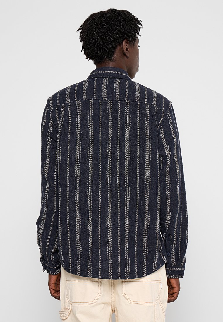 Marineblauwe shirt met verticale witte streepjes, lange mouwen en een kraag. De stof lijkt textuur te hebben met een casual, ontspannen pasvorm.