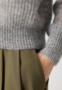 Grauer Strickpullover mit gerippter Textur, kombiniert mit einem olivgrünen, plissierten Stoff. Sichtbar ist eine Hand, die in die Tasche der Hose gesteckt ist.