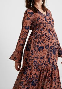 Gravid kvinna i en långärmad, rostfärgad klänning med marinblå blommönster och trumpetsärmar.