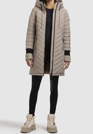 Femme portant un long manteau matelassé beige avec capuche, des leggings noirs et des bottes beiges à lacets, debout devant un fond uni.