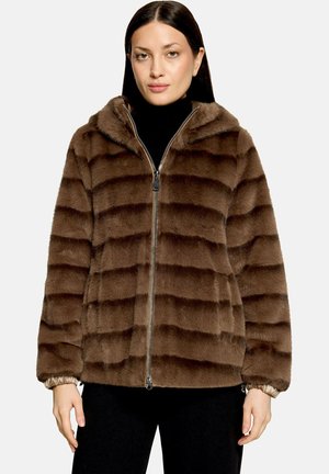Vrouw draagt een bruine gestreepte faux fur jas met rits en capuchon en zwarte broek tegen een effen achtergrond.