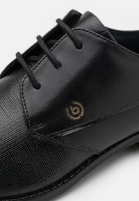 Scarpa in pelle nera con finitura texturizzata, dotata di lacci tondi, logo dorato e design della punta liscia.
