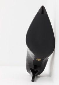 Chaussure à talon haut noire à bout pointu vue de la semelle, avec un petit logo doré près du talon, reposant contre une surface blanche.