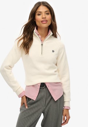 Femme avec de longs cheveux portant un pull crème à col zippé quart sur une chemise rose et un pantalon gris à rayures noires avec une ceinture noire, posant avec une main dans la poche.